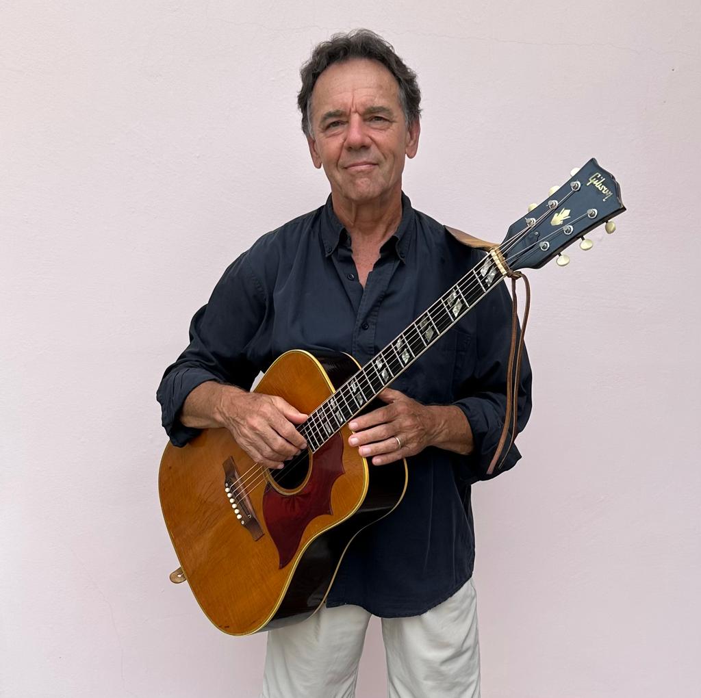 A man holding an acoustic guitar in front of a white wall.